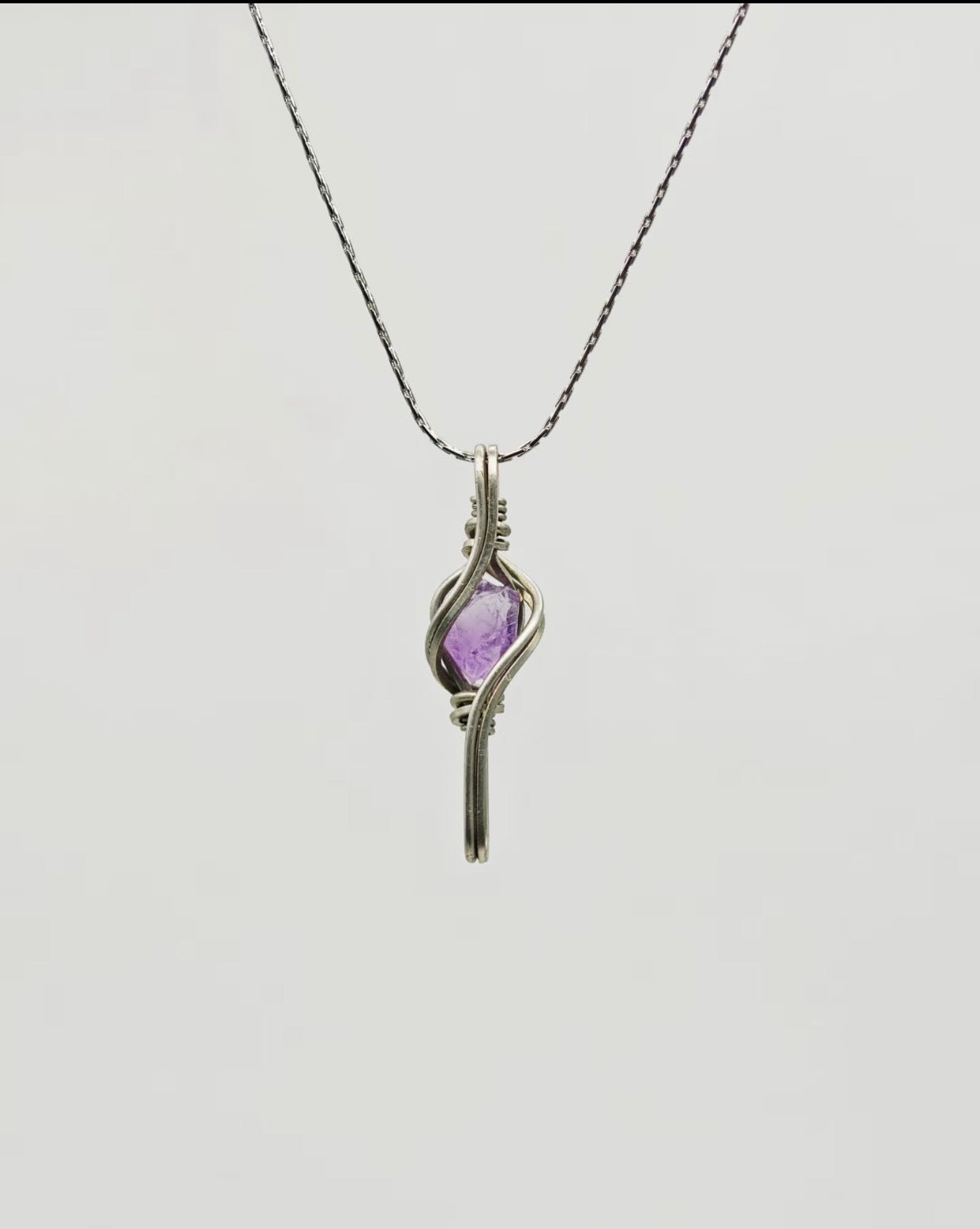 Purple crystal pendant framed by elongated flowing metal lines, suspended from a chain on a light background.