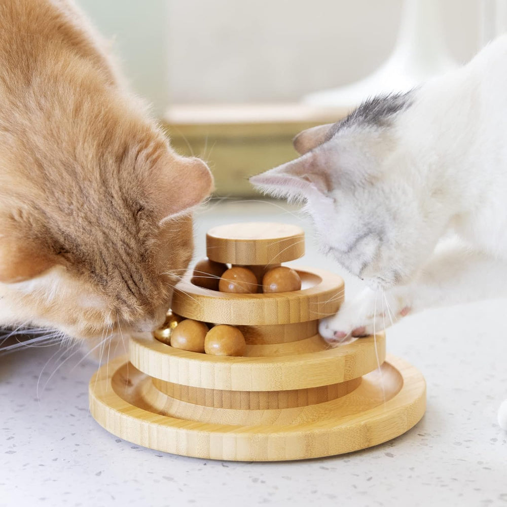 Two cats looking at a tiered ball-track tower with wooden balls on a light surface.