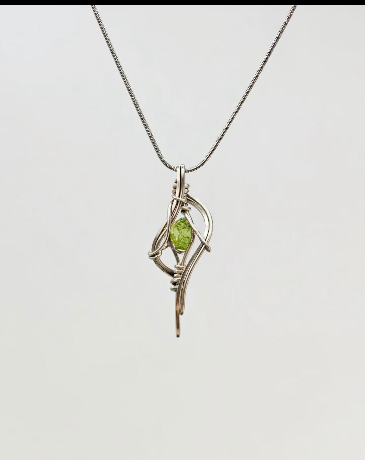 Green crystal pendant enclosed in curved metal lines, suspended from a chain against a light background.