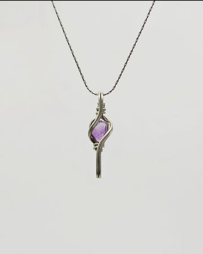 Purple crystal pendant framed by elongated flowing metal lines, suspended from a chain on a light background.